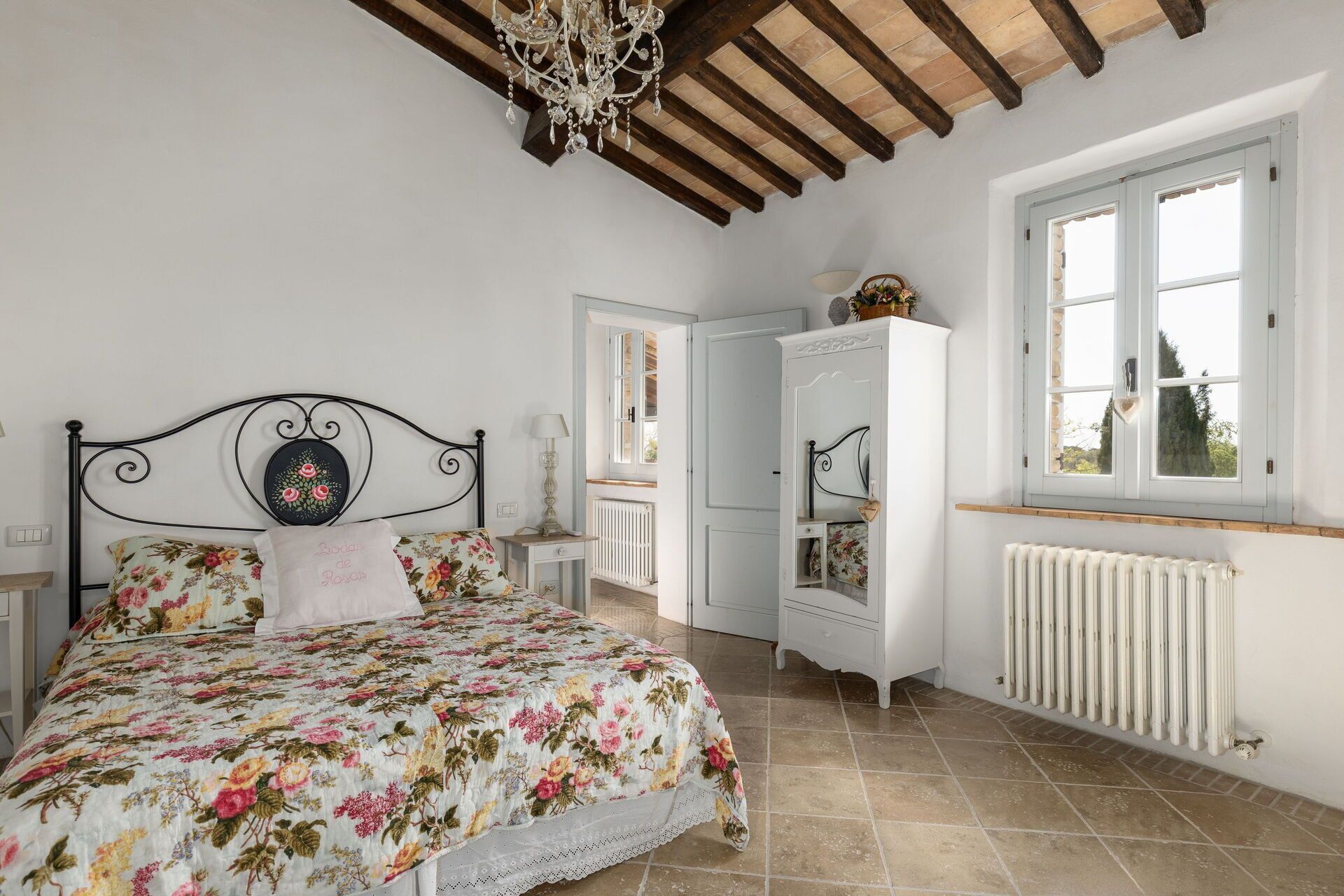 A bedroom with a floral bedspread, a white wardrobe, a radiator, and a window with a view.