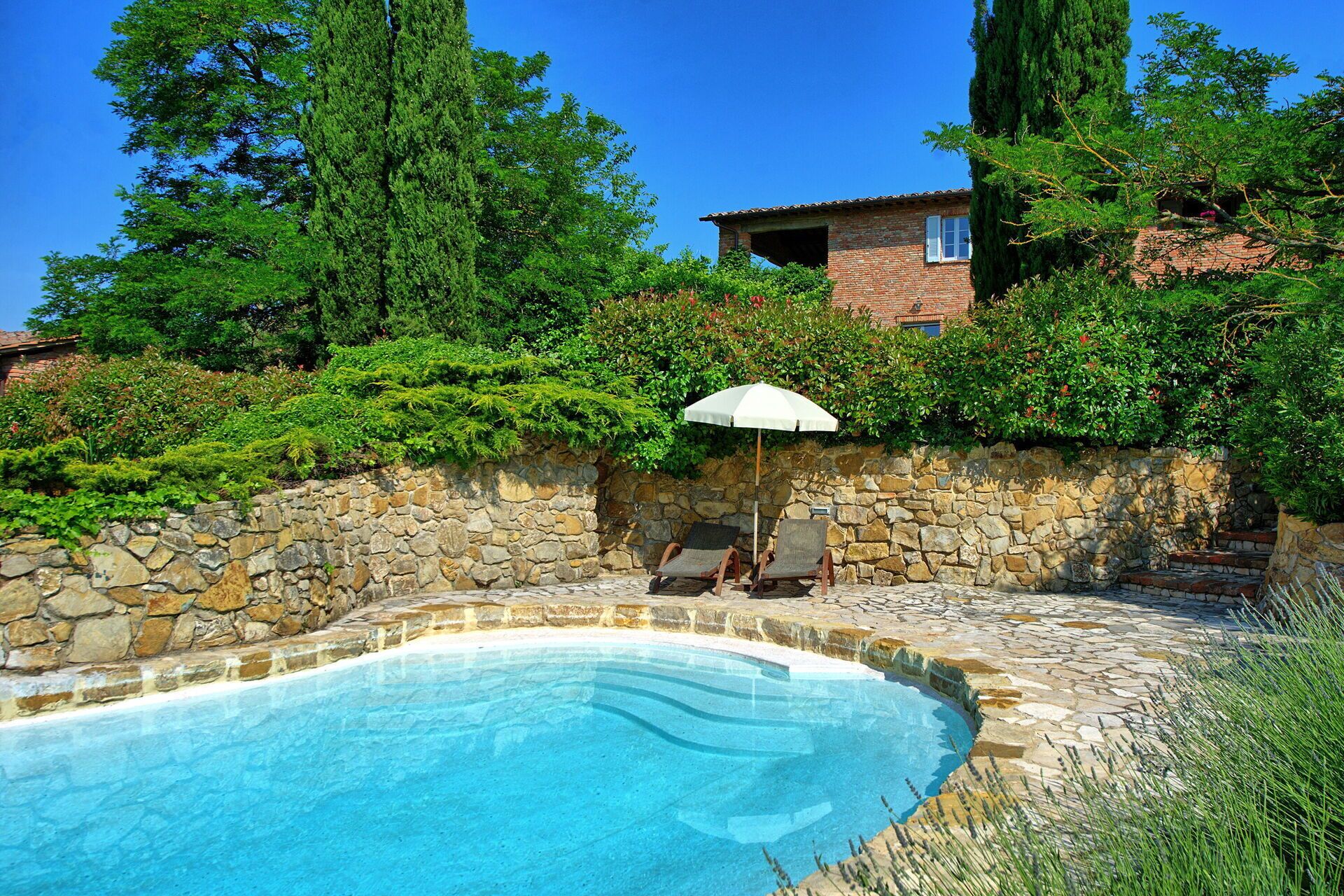 A swimming pool with stone walls, a stone pathway, and two lounge chairs with an umbrella.