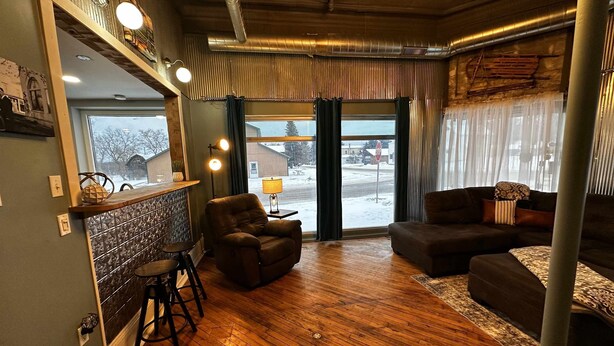 Ein gemütliches Zimmer mit Holzboden, einer Couch, einem Sessel, einem kleinen Tisch mit Lampe und einem Fenster mit Vorhängen.