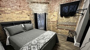 Ein Schlafzimmer mit Backsteinwand, einem Bett mit gemusterten Bettwaren, einem Fenster mit schwarzem Vorhang und einem kleinen schwarzen Stuhl.