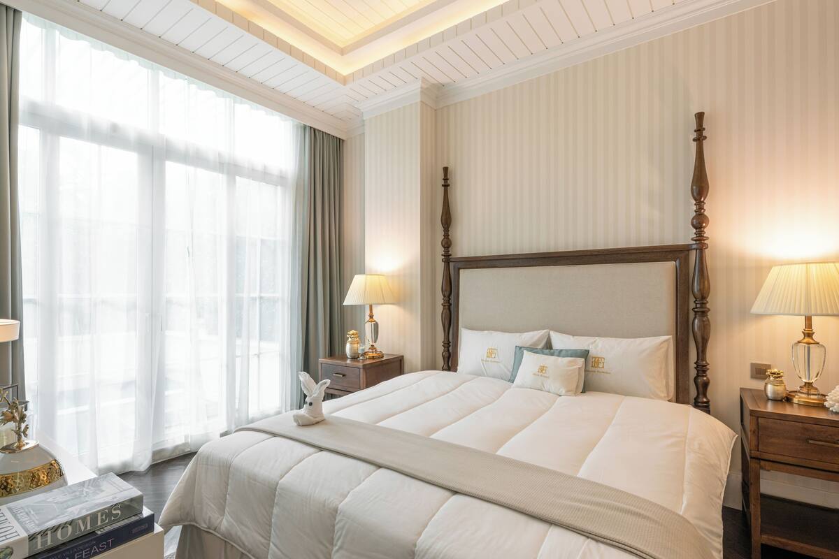 A large bed with a wooden headboard, two bedside tables with lamps, a window with sheer curtains, and a decorative rabbit figurine on the bed.