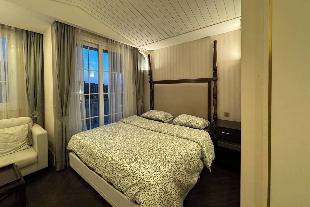 A hotel room with a large bed, a bedside table, a chair, and a window with curtains.
