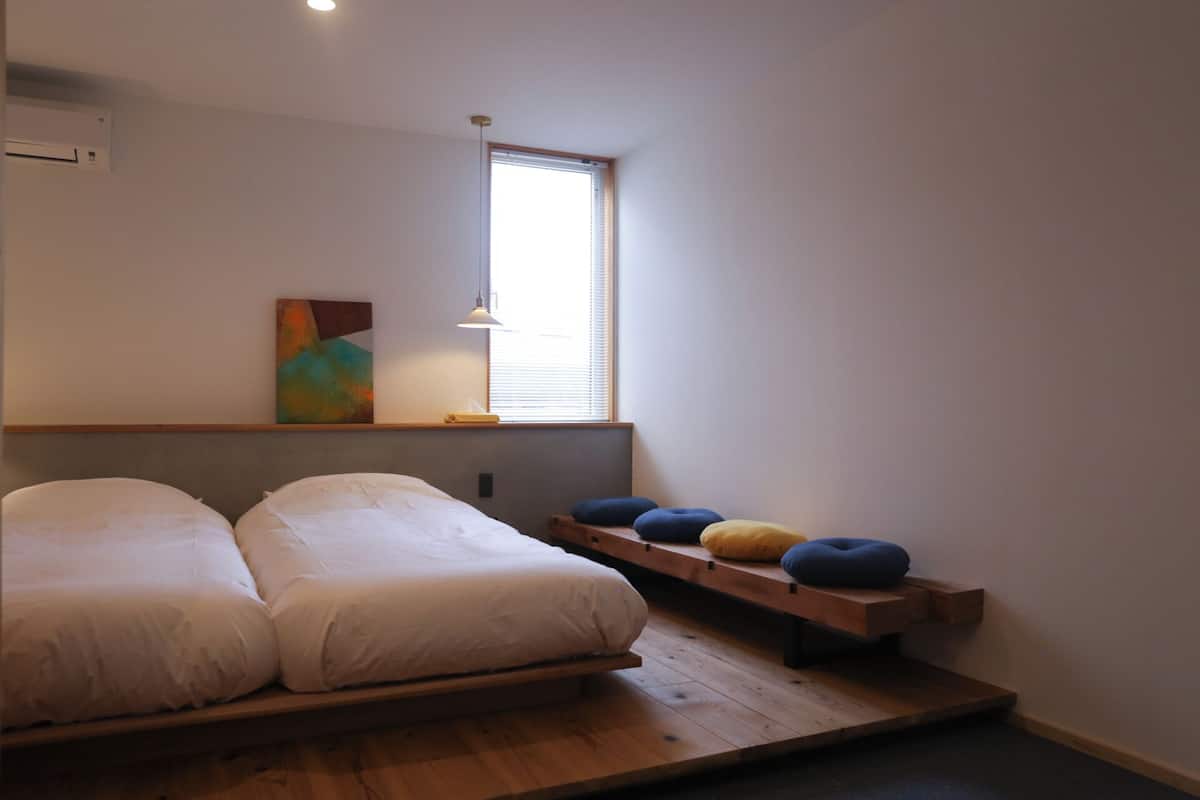 Ein schlichter Schlafzimmer mit zwei Betten, einem Fenster und einer Holzplattform mit Kissen.