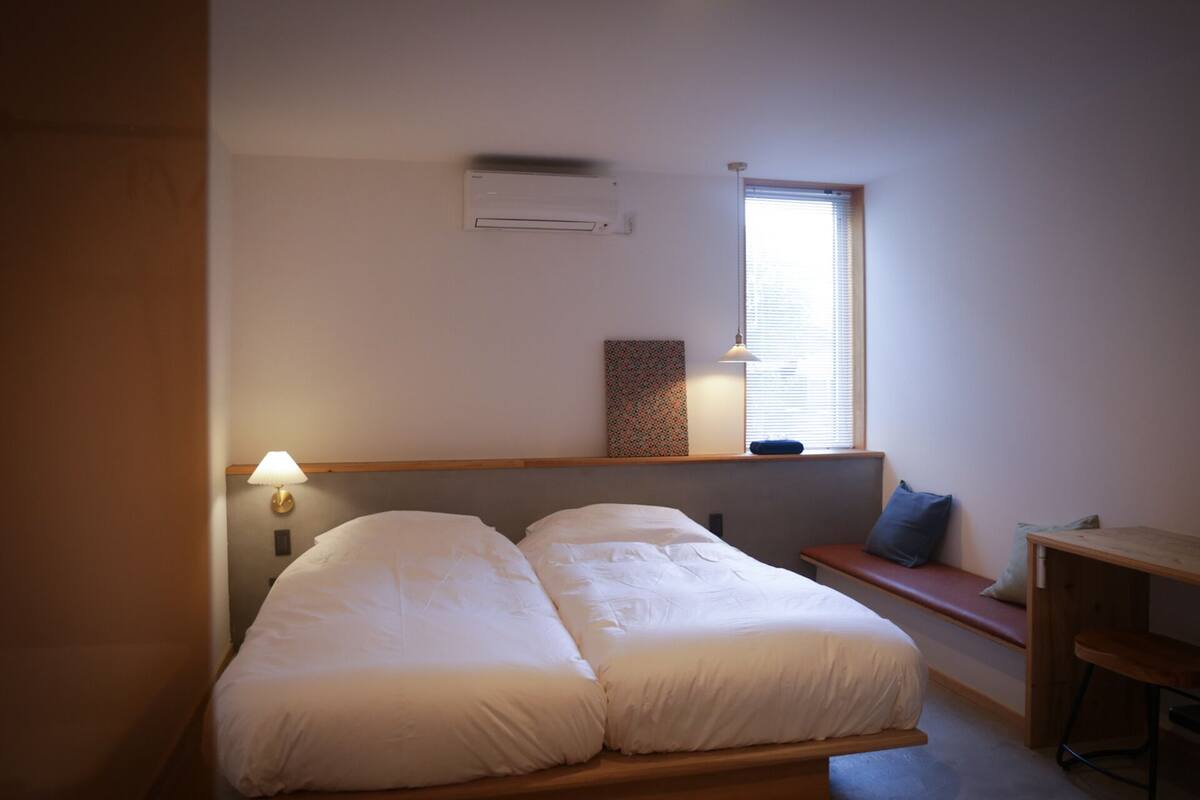Ein Schlafzimmer mit Bett, Nachttisch, Lampe, Klimaanlage, Fenster und kleinem Schreibtisch.