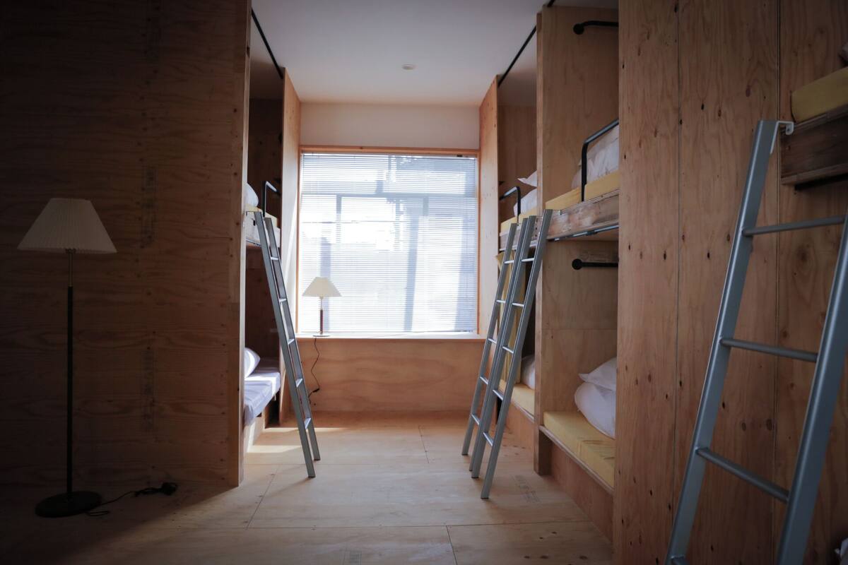 Ein schmaler Flur mit Etagenbetten und Leitern, Holzwänden und einem Fenster mit Jalousien.