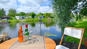 A wooden table with a bottle, glasses, and a placemat by a calm lake with houses in the background. - Holiday Home 'Reiseoase' with Lake View, Wi-Fi and Air Conditioning (Bützow)
