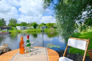 Une table en bois, avec une bouteille, des verres et un set de table, placée au bord d’un lac paisible, avec des maisons en arrière-plan.