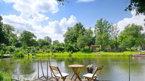 A wooden deck with two chairs and a small table, overlooking a calm pond with a boat. - Holiday Home 'Reiseoase' with Lake View, Wi-Fi and Air Conditioning (Bützow)