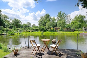 Een houten terras met twee stoelen en een kleine tafel, met uitzicht op een rustig meer met een boot.