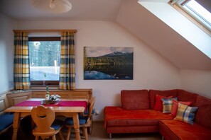 Ein Zimmer mit Esstisch, Stühlen, einem roten Sofa, einem Fenster mit Vorhängen und einem Gemälde einer Berglandschaft.
