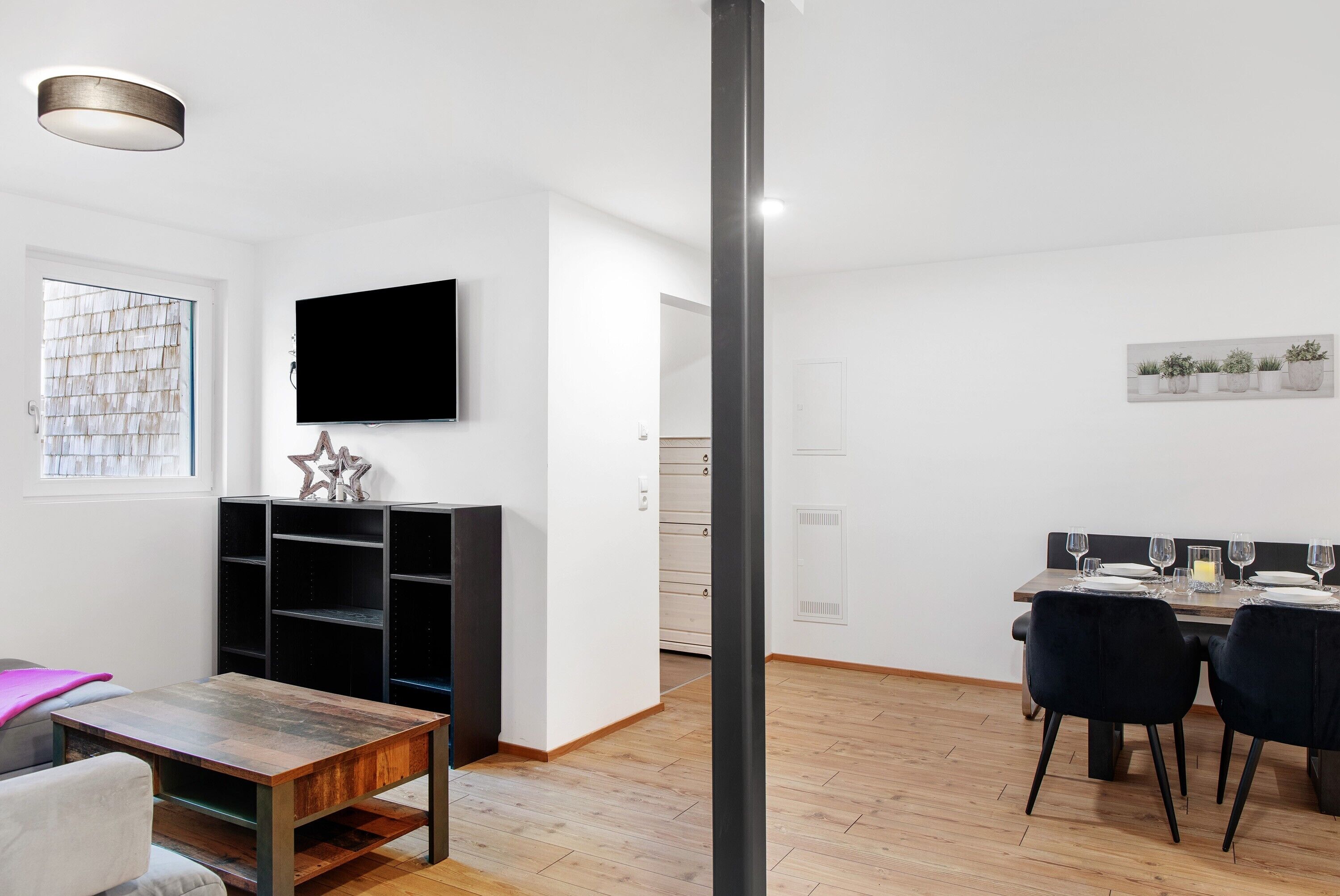 A modern living room with a wooden coffee table, black chairs, a flat-screen TV, and a black shelving unit.