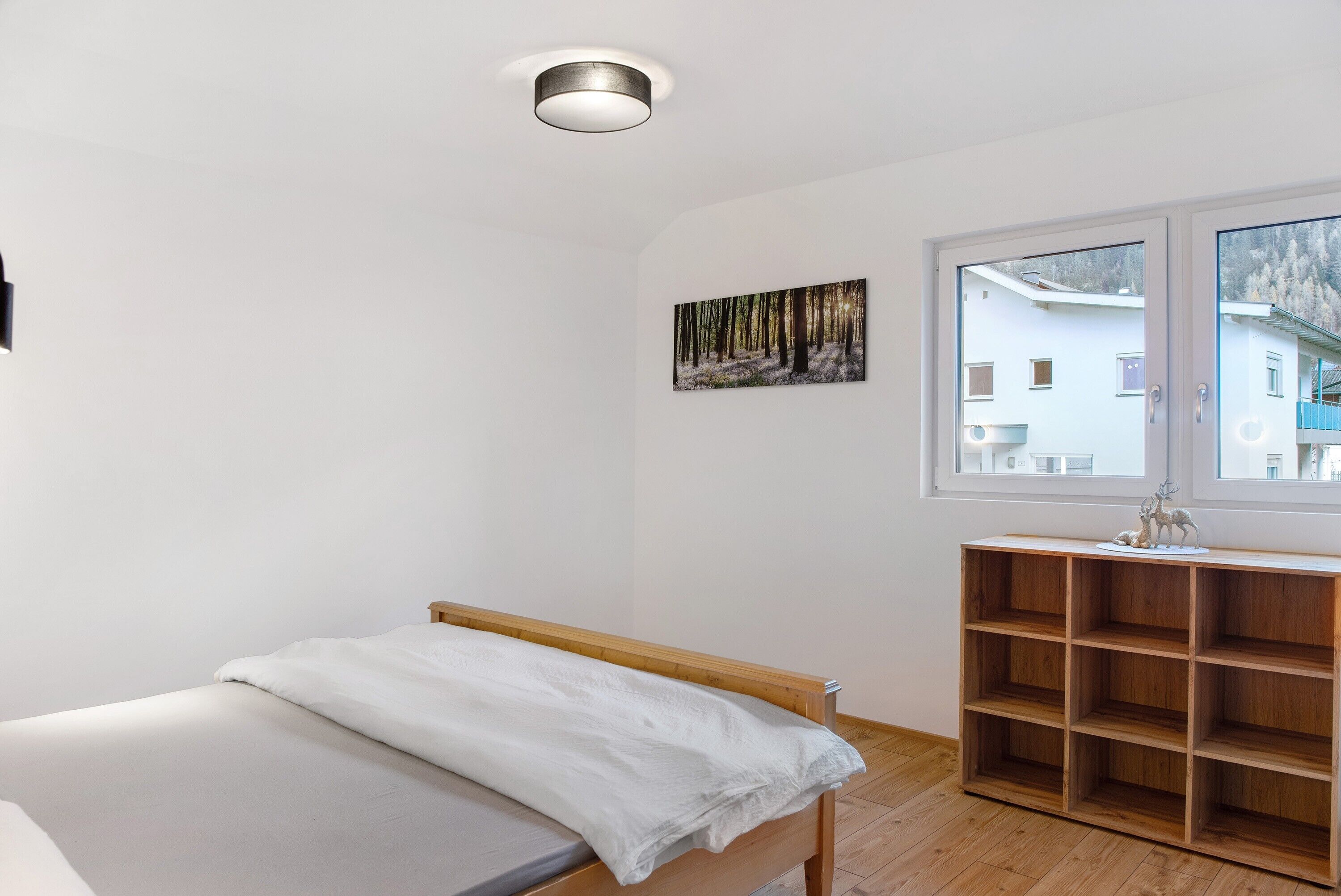 A bedroom with a bed, a wooden shelf unit, a window with a view of buildings, and a ceiling light fixture.
