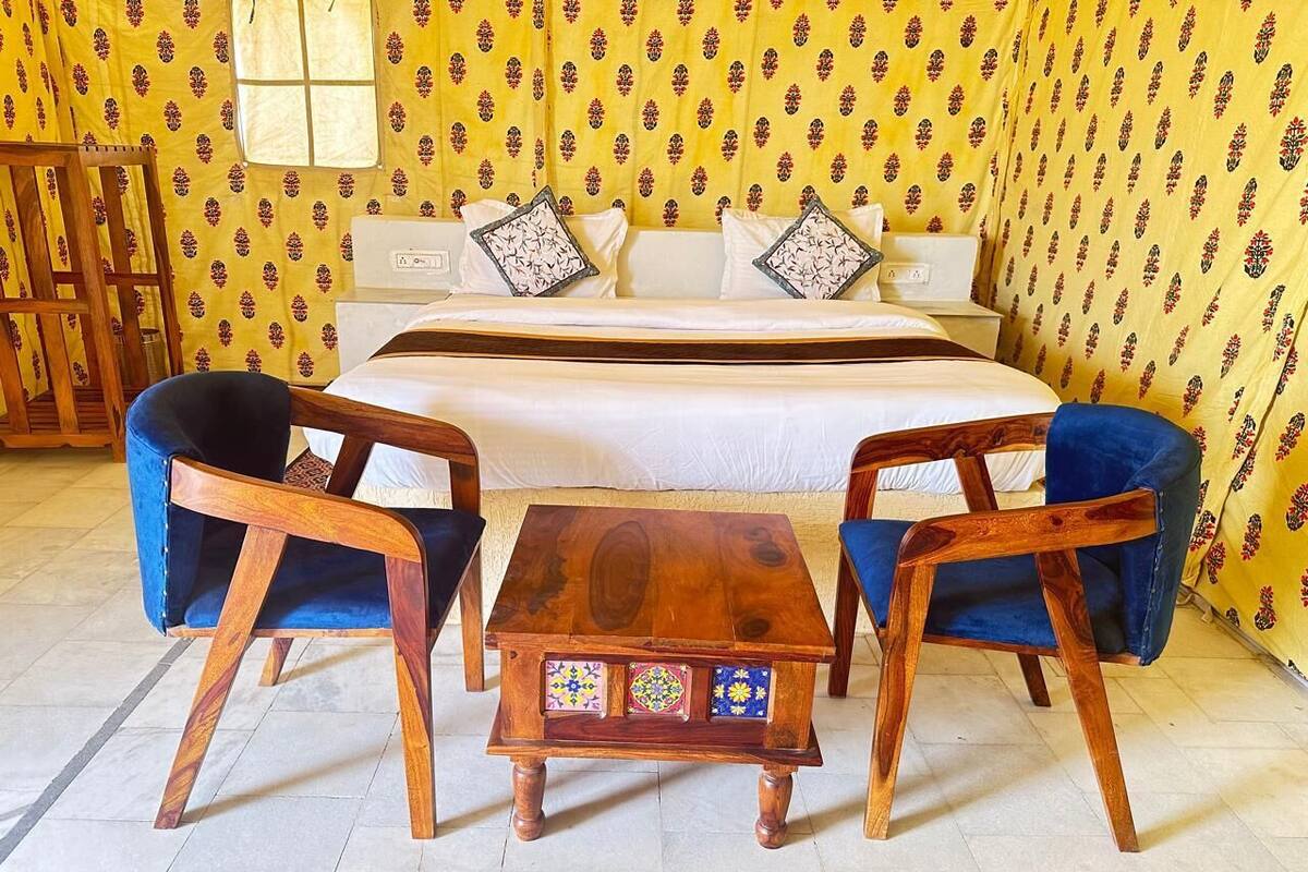 A room with a bed, two blue chairs, a wooden table, and patterned yellow wallpaper.