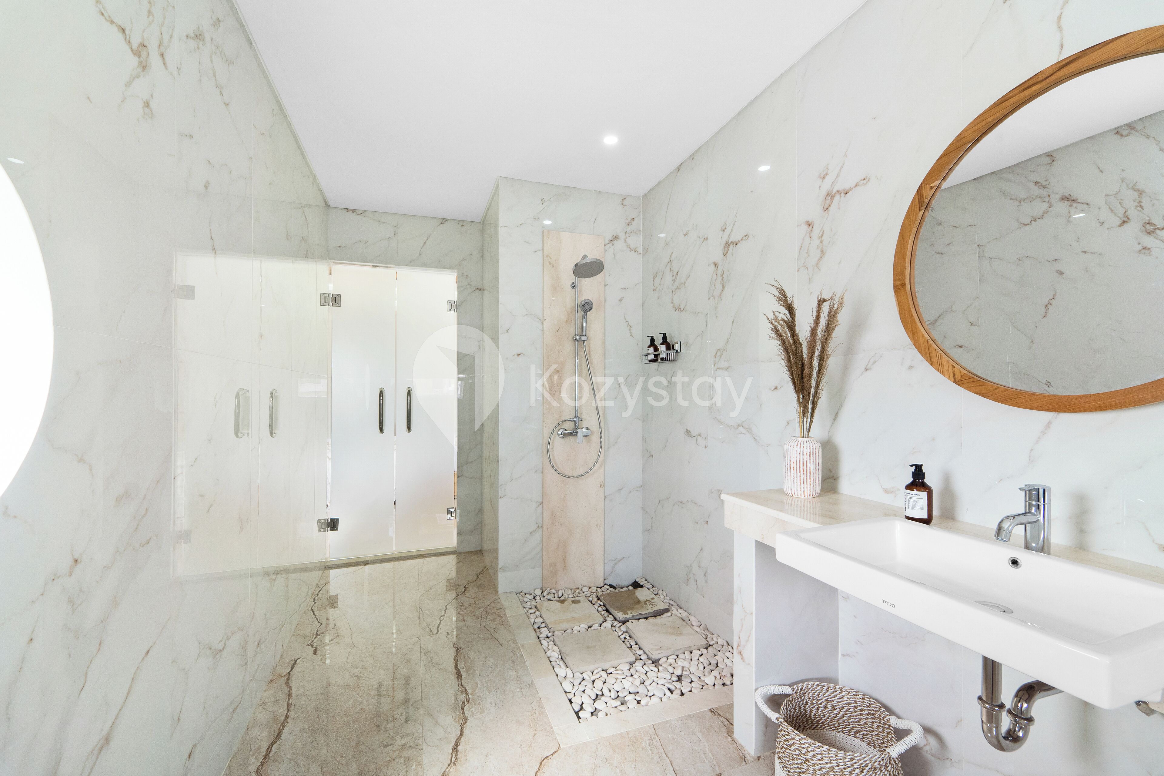 A modern bathroom with a marble floor, a shower with a wooden panel, a white sink with a mirror, and a small vase with dried plants.