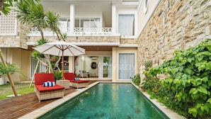 Zona de piscina con sillones de mimbre, terraza de madera y pared de piedra.