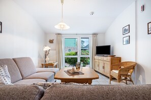 Un salon comprenant un canapé, une table basse en bois, un fauteuil en osier, un téléviseur et une fenêtre donnant sur un paysage verdoyant.