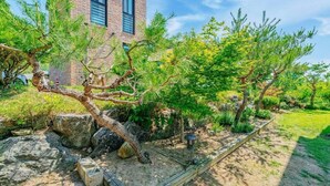 A landscaped garden with a variety of trees, rocks, and a brick building in the background. - Chuncheon Stay 266 (Chuncheon)