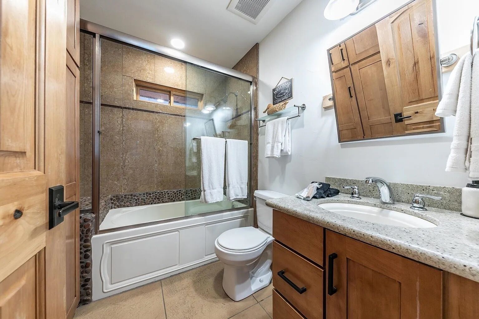 Ein modernes Badezimmer mit einer gläsernen Dusche, einer weißen Toilette, Holzschränken und einem Waschbecken mit Granitplatte.