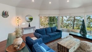 A modern living room with a blue sofa, a wooden coffee table, and a large window offering a view of the outdoors. - Spacious 4-Bed Home with Coastal Charm (Long Beach)