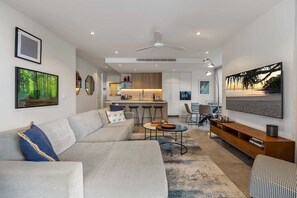 A modern living room with a sofa, coffee table, and TV. There is a kitchen area with a bar and stools.