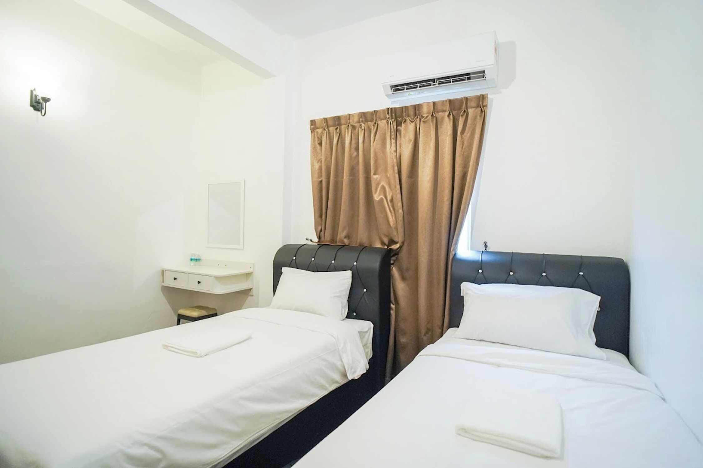 A hotel room with two single beds, a small bedside table with a lamp, and a window with brown curtains.