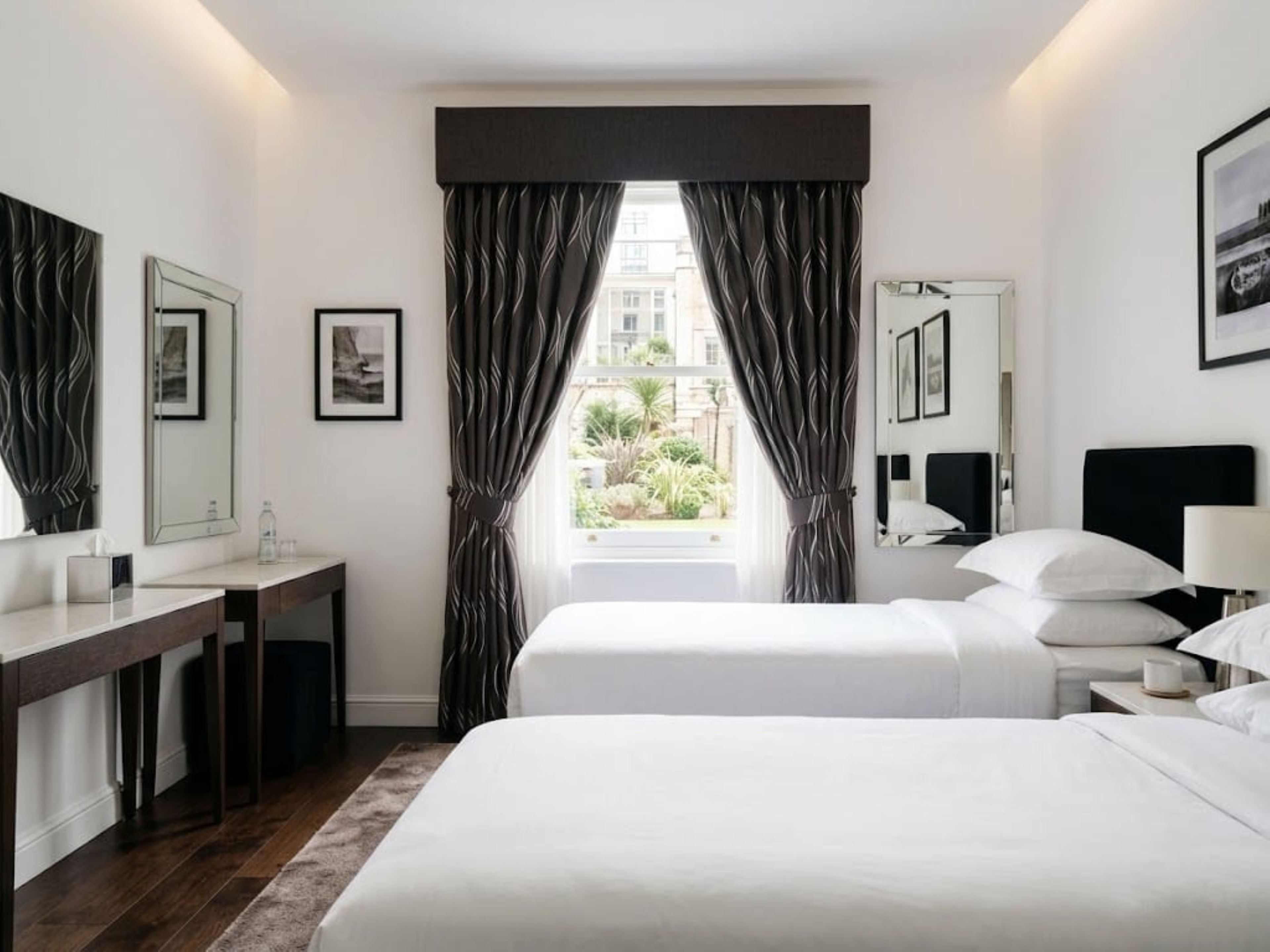 Hotellihuone, jossa on kaksi sänkyä, työpöytä, peili ja ikkuna, jossa on verhot.