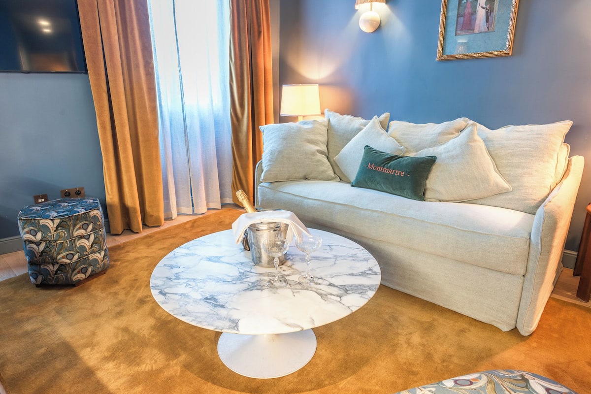 A room with a white sofa, a marble coffee table, a patterned ottoman, and a lamp.