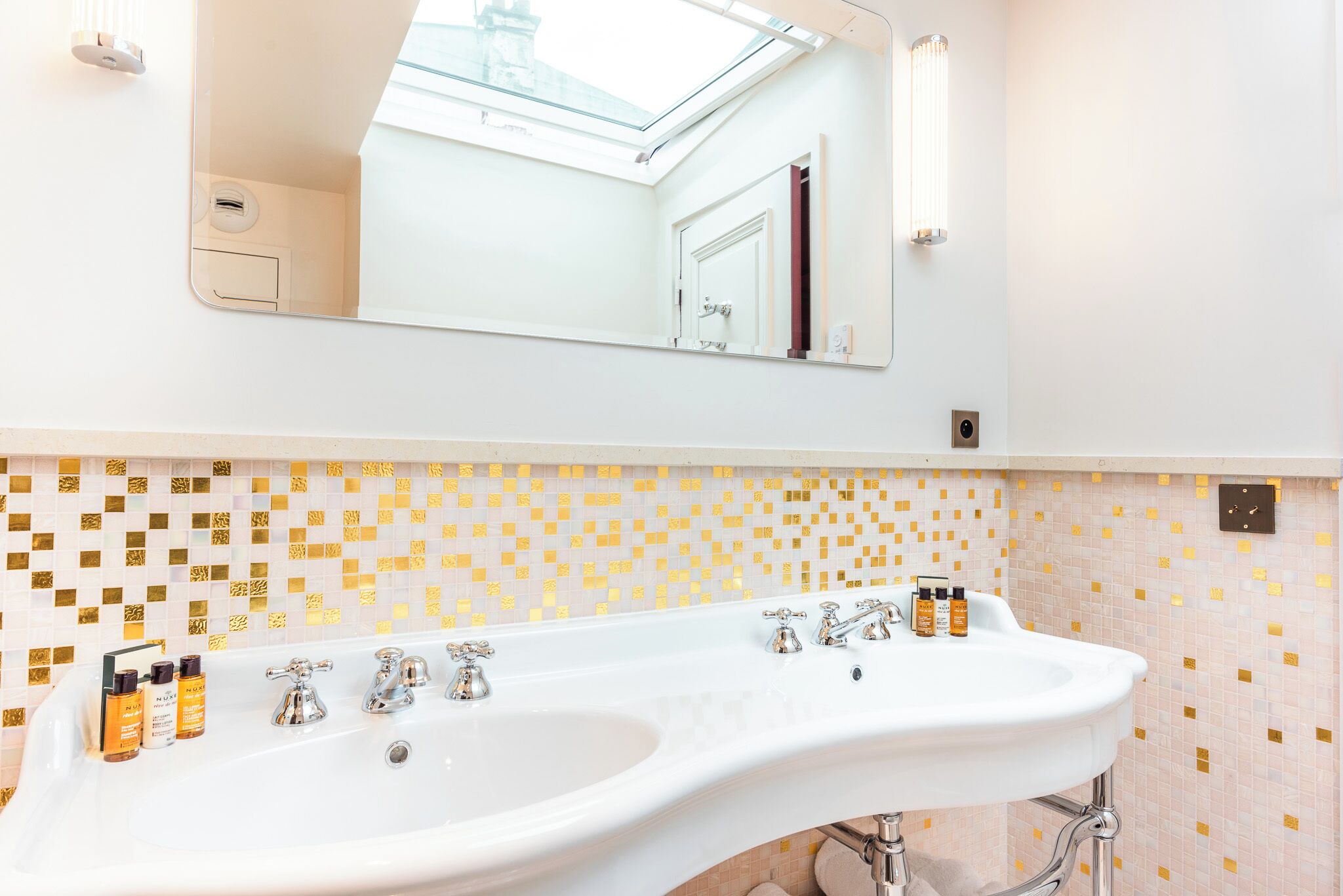 A bathroom with a white sink, gold mosaic tiles, a large mirror, and a skylight.