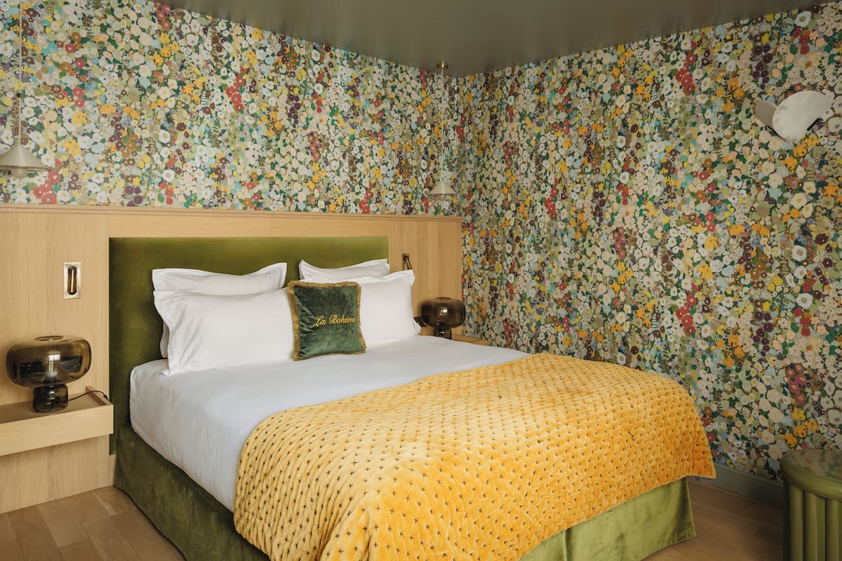 A room with a floral wallpaper, a green upholstered headboard, a bed with white linens and a yellow blanket, and a nightstand with a lamp.