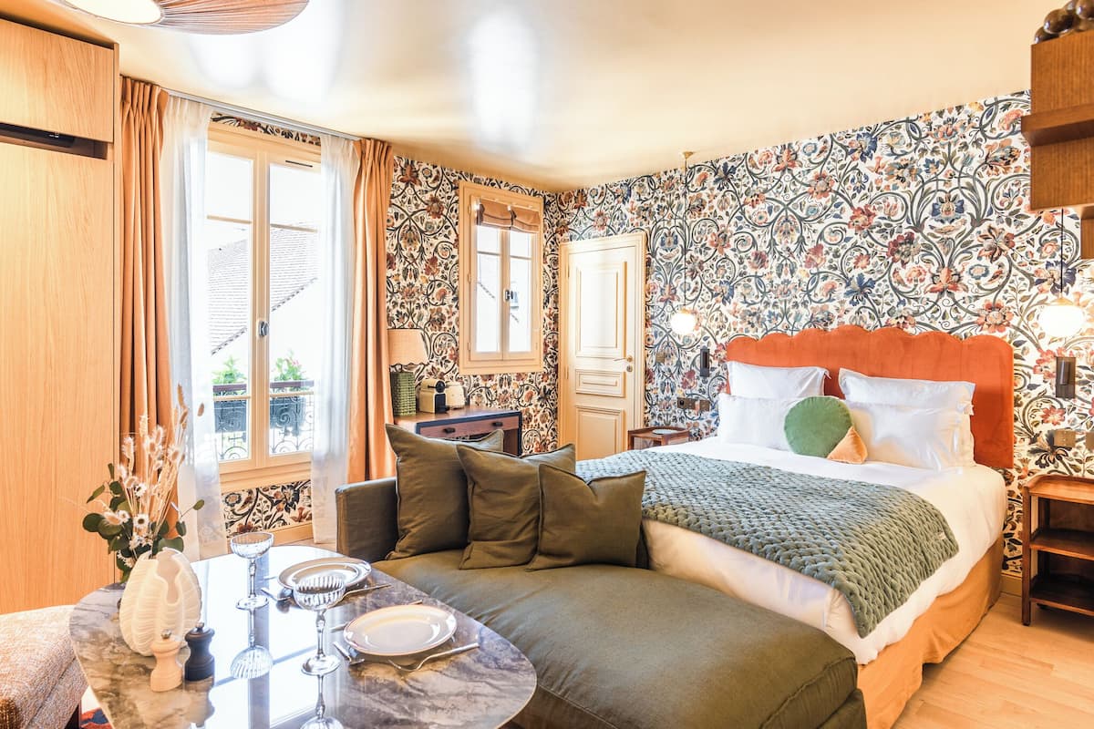 A bedroom with a bed, a dining table with chairs, a vase with flowers, and a window with curtains.