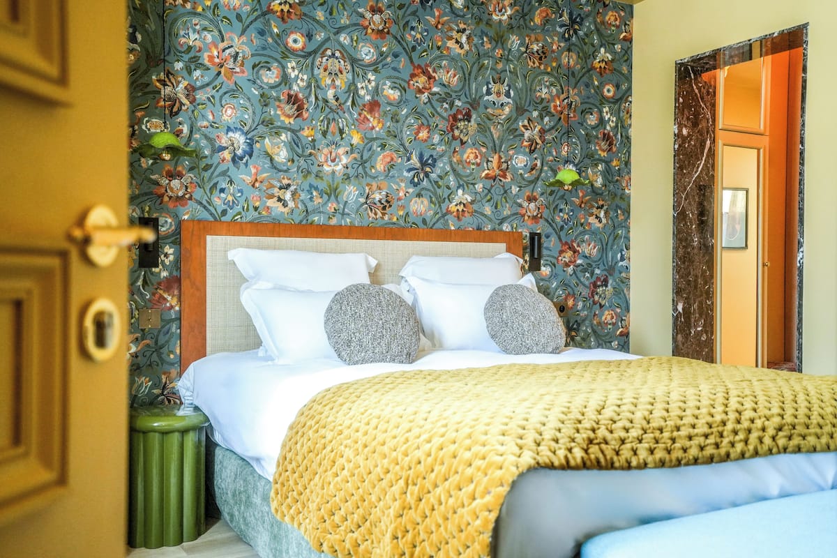 A hotel room with a floral wallpaper, a bed with white linens and two pillows, a green trash bin, and a wooden door.