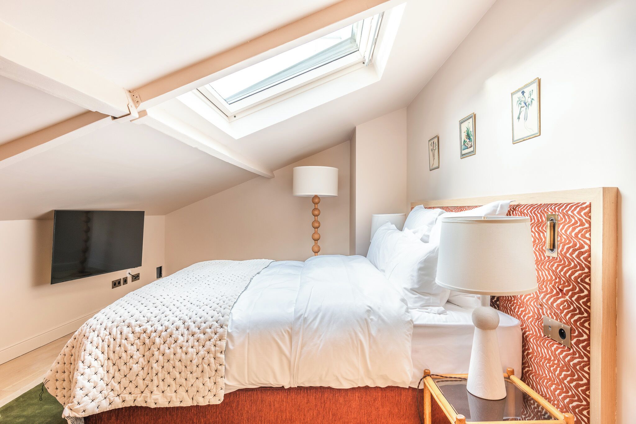 A bedroom with a bed, a bedside table, a lamp, a TV, and a wall with framed pictures.