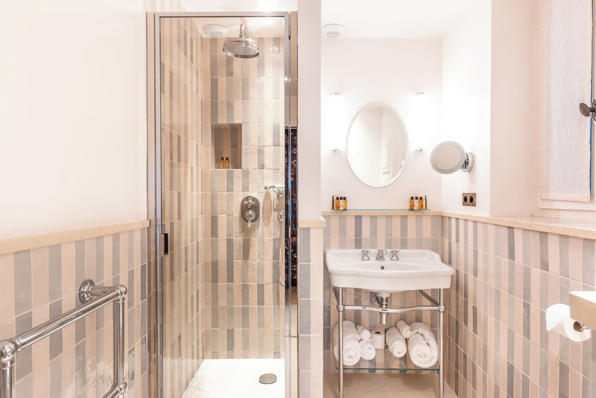 Une salle de bain moderne avec une douche à parois de verre, un lavabo blanc doté d’une étagère en verre et des murs recouverts de carreaux à rayures.