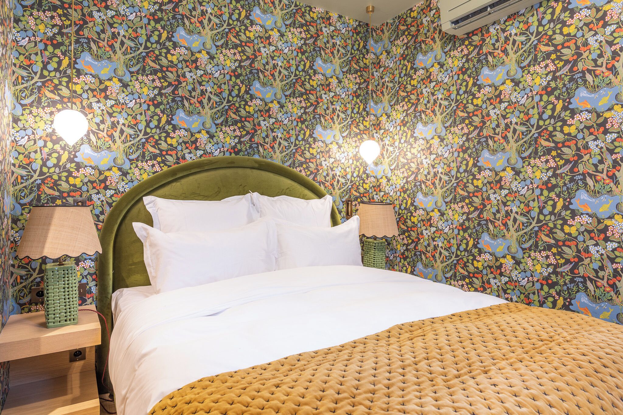 A bed with white bedding, a green headboard, and a woven throw blanket. There are two bedside lamps on wooden nightstands. The walls are covered with a floral wallpaper.