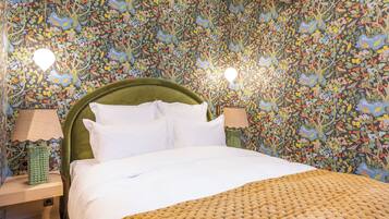 A bed with white bedding, a green headboard, and a woven throw blanket. There are two bedside lamps on wooden nightstands. The walls are covered with a floral wallpaper.