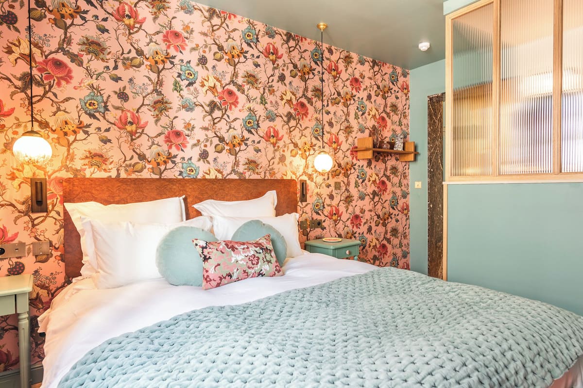 A bedroom with floral wallpaper, a wooden headboard, a blue knitted bedspread, and two bedside lamps.
