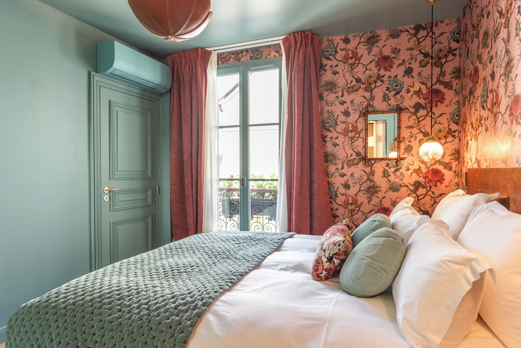 A bedroom with a bed, floral wallpaper, a window with curtains, and a door.