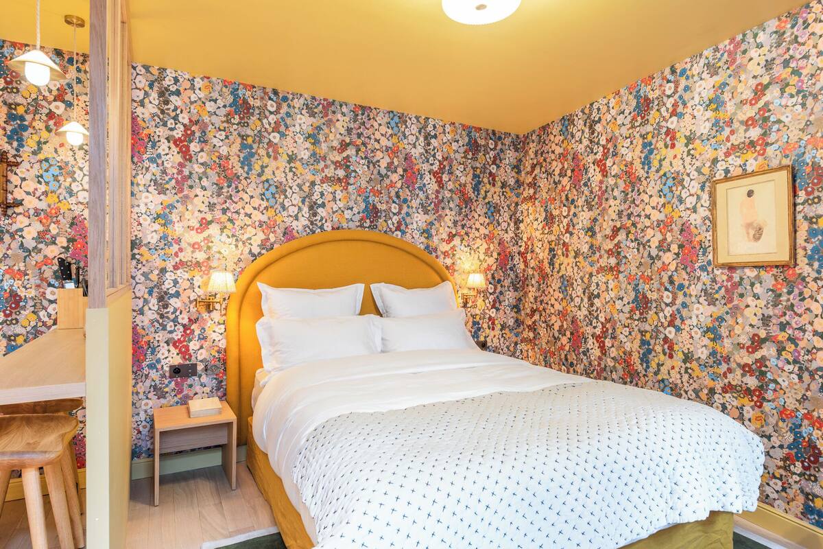 A bedroom with a floral wallpaper, a wooden headboard, a bedside table with a lamp, and a framed picture on the wall.