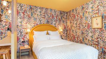 Ein Schlafzimmer mit floralem Wandpapier, einem hölzernen Kopfteil, einem Nachttisch mit Lampe und einem gerahmten Bild an der Wand.