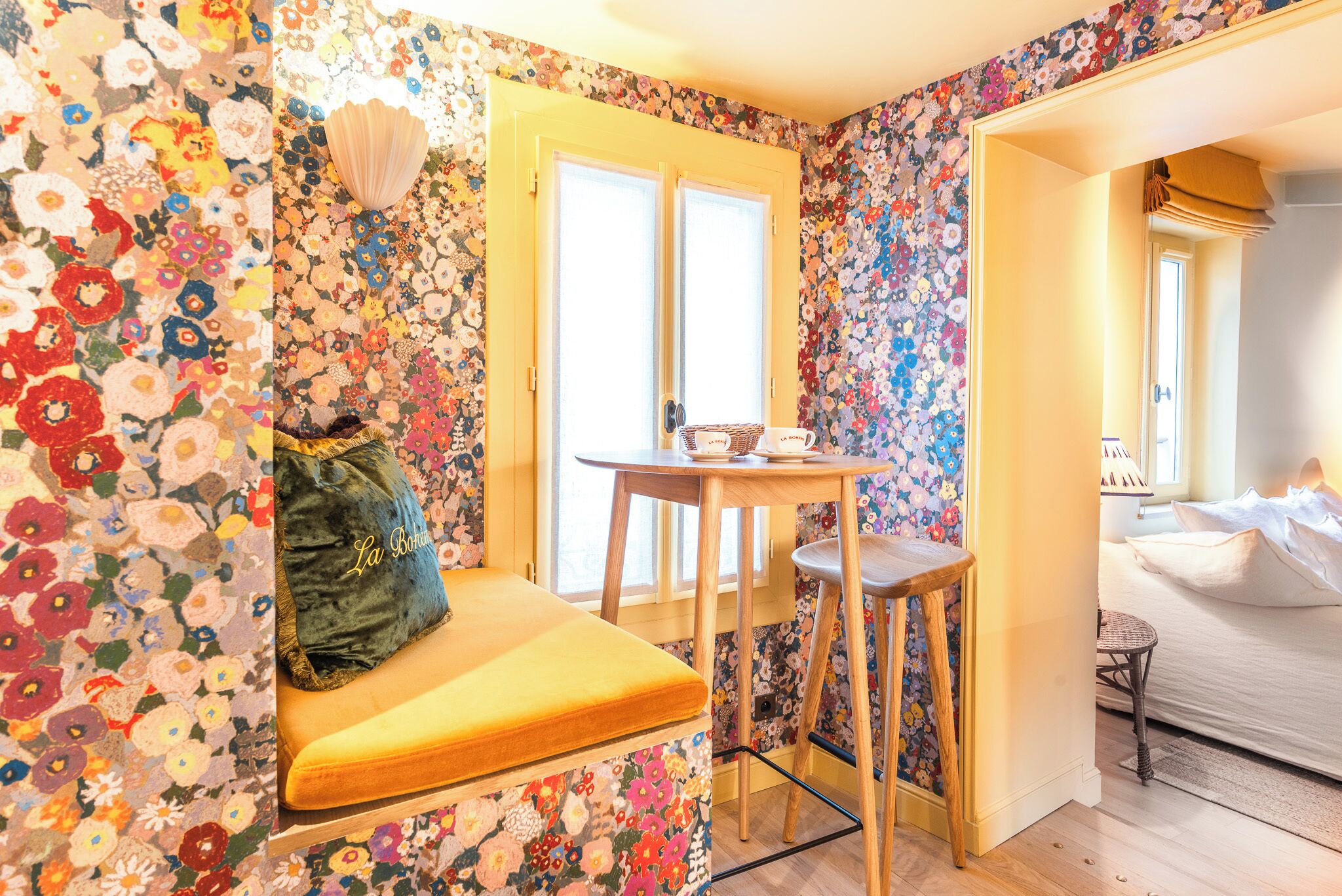 Une pièce avec un papier peint à motifs floraux, un banc jaune, une petite table en bois et un lit recouvert de draps blancs.
