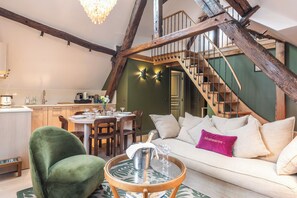 A cozy living room with a green armchair, a white sofa with a 'Montmartre' pillow, and a wooden coffee table. A staircase with exposed beams leads to a dining area with a table set for four. - Maison La Bohème Montmartre - Studios & Apartments (Paris)
