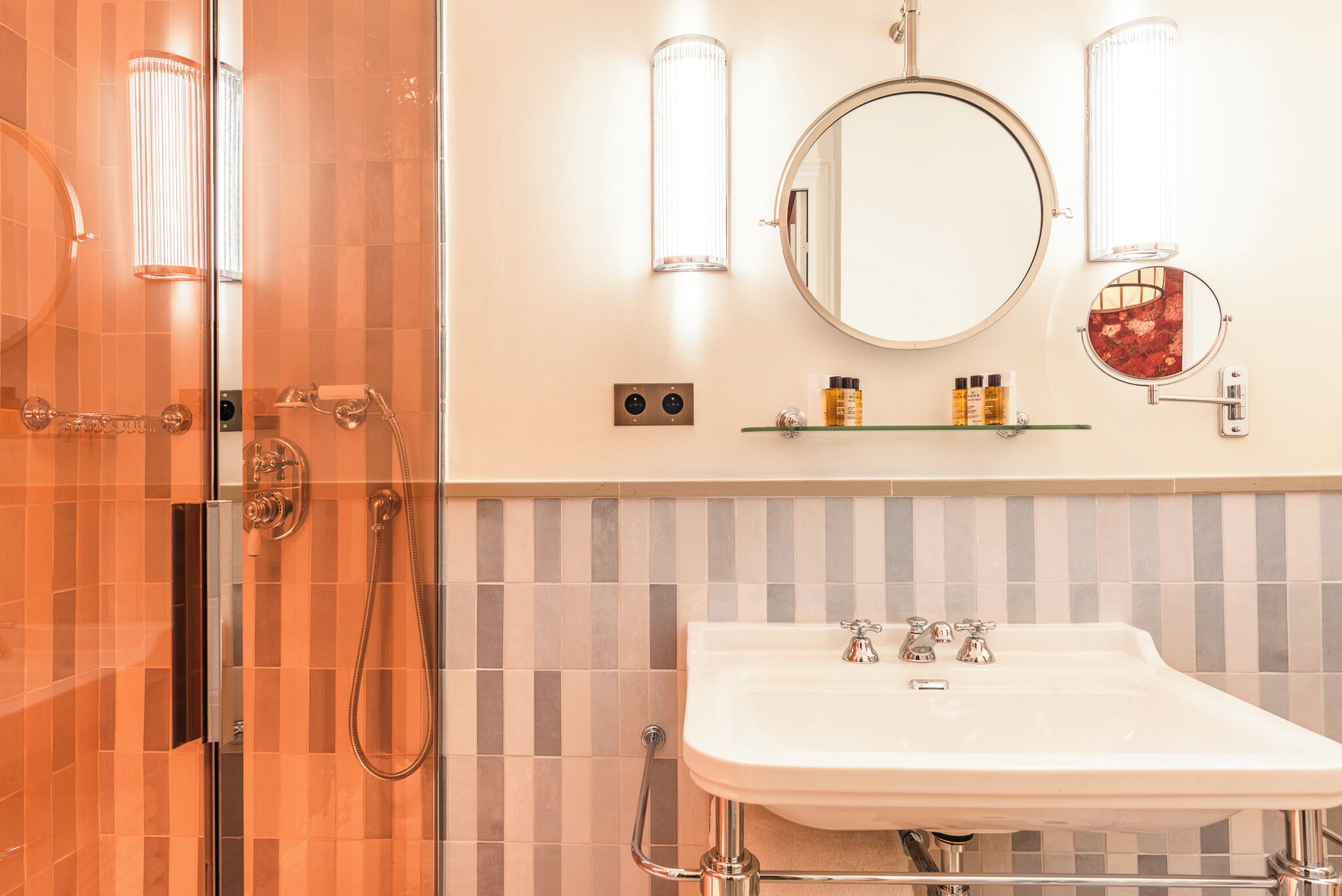 Un bagno con box doccia in vetro, lavandino bianco con due rubinetti e specchio rotondo sopra il lavandino.