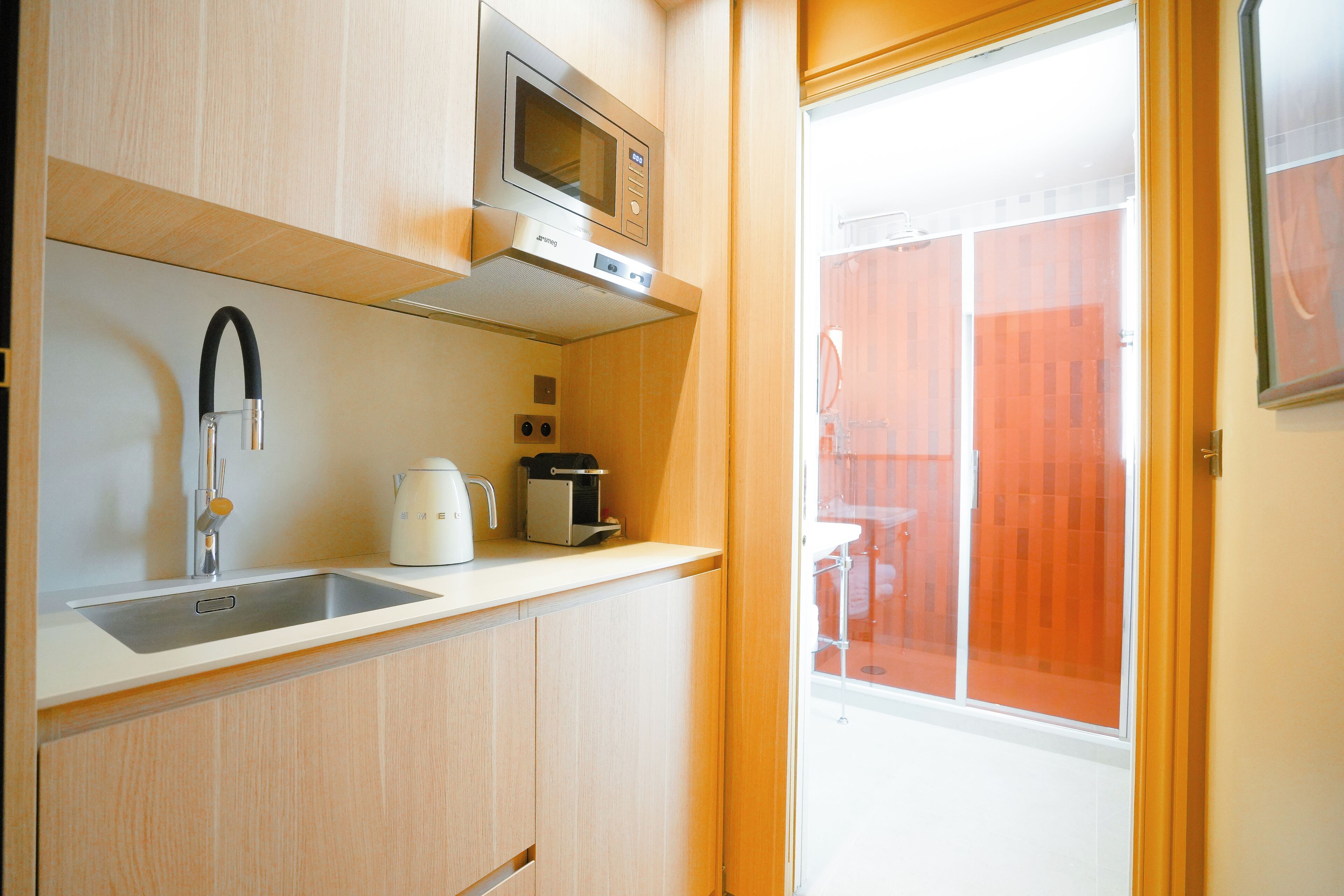 A compact kitchen with a microwave, kettle, and coffee maker on the counter, and a window with a view of a red fence.