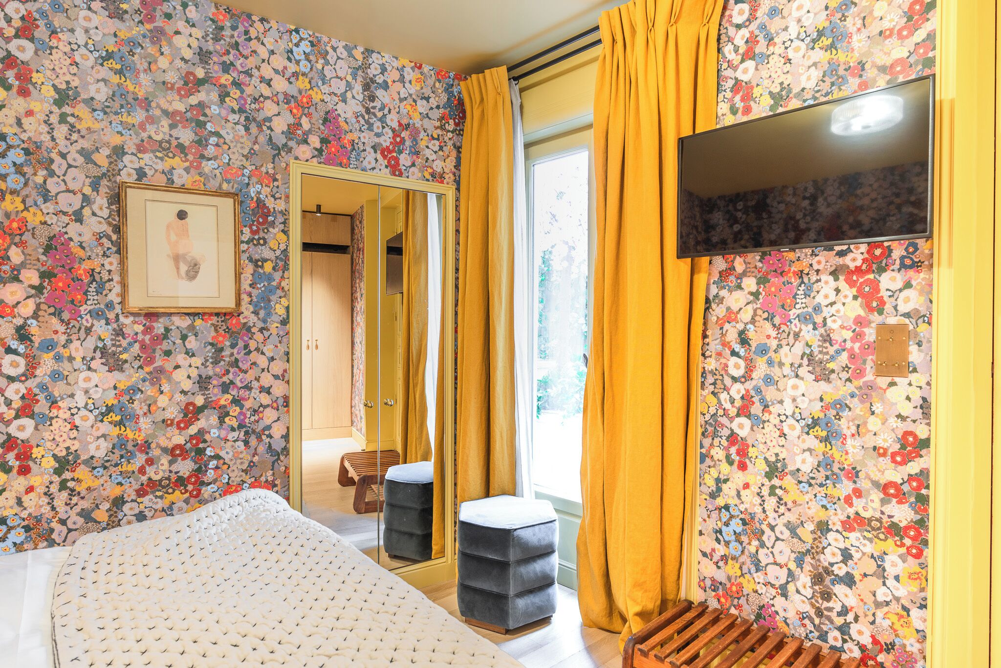 A bedroom with floral wallpaper, a bed, a mirror, a television, and a window with curtains.