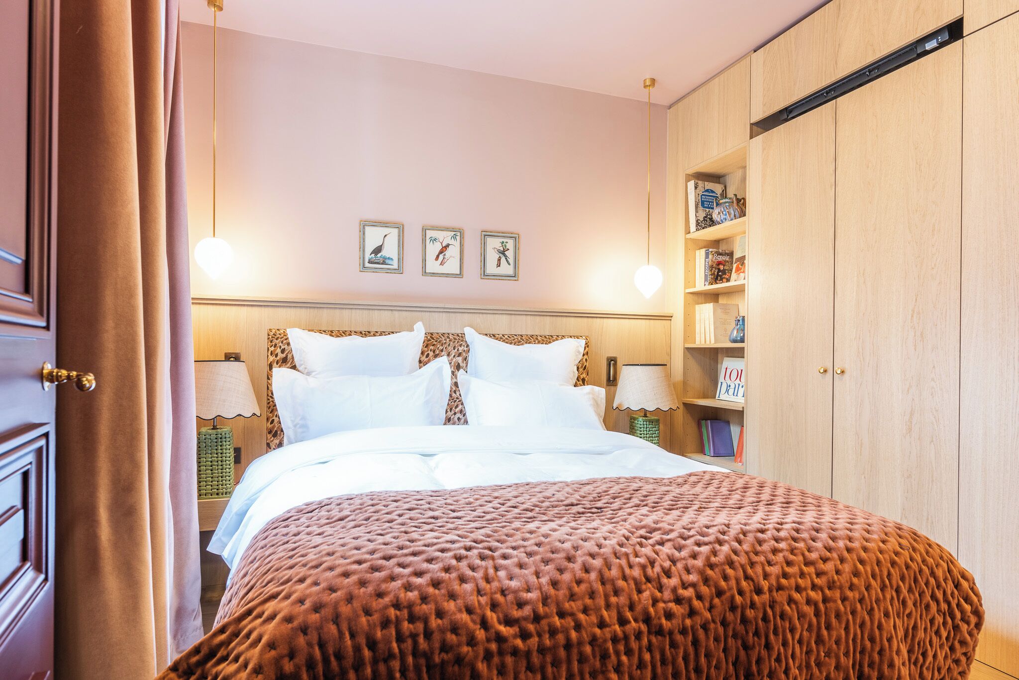 A neatly made bed with a brown textured blanket, a headboard with wall art, bedside lamps, and a wooden wardrobe with shelves.