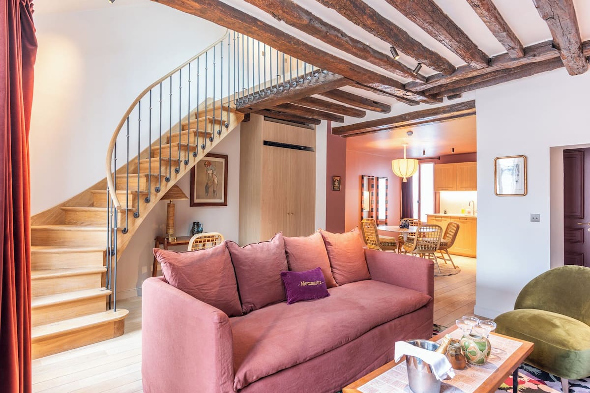 A living room with a wooden staircase, a sofa, a dining area, and a kitchen in the background.