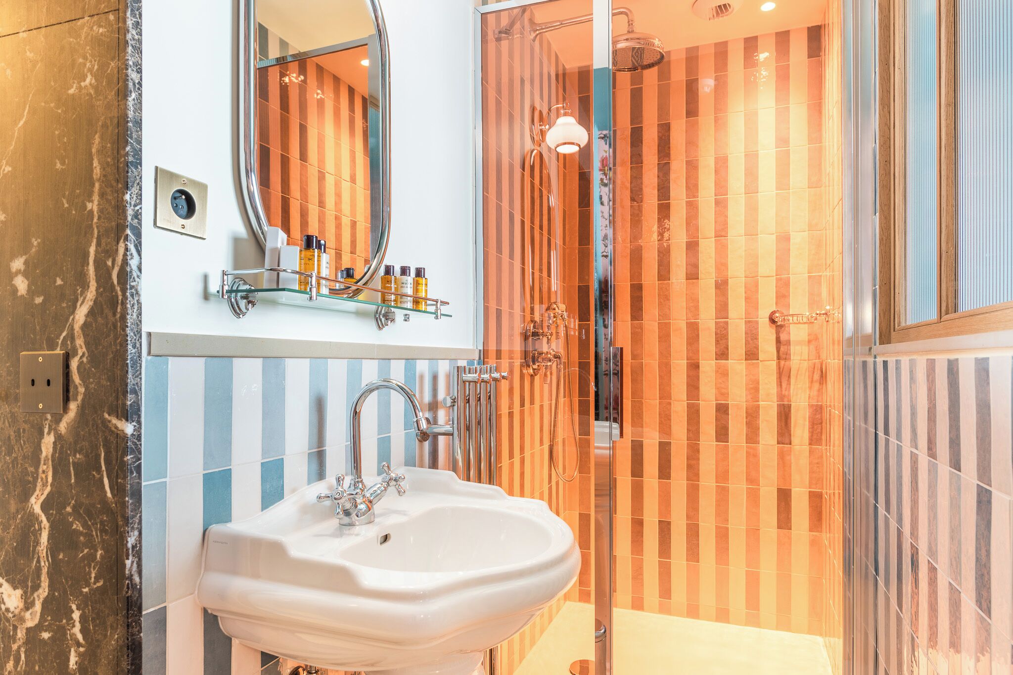 Un baño con lavamanos blanco, un espejo y una ducha con azulejos anaranjados.