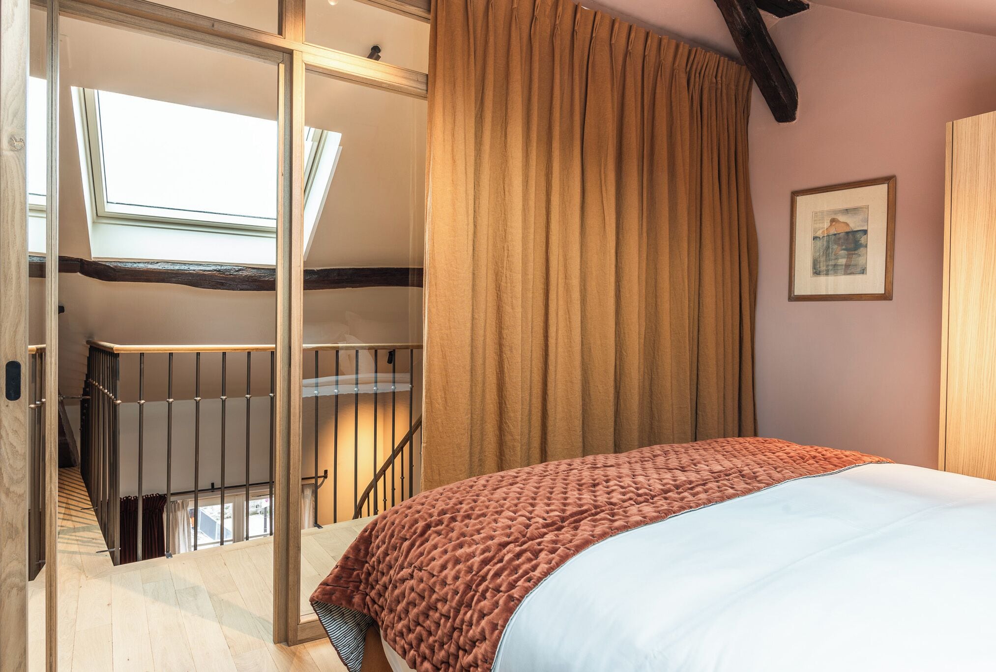 A bedroom with a bed, a large window, a staircase, and a painting on the wall.