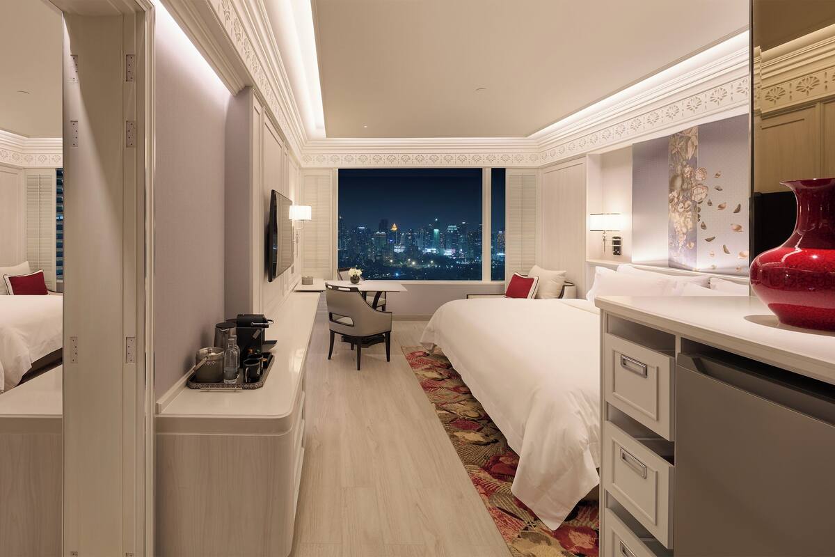Une chambre d’hôtel avec deux lits, un bureau, une chaise, une grande fenêtre donnant sur la ville et un vase rouge.