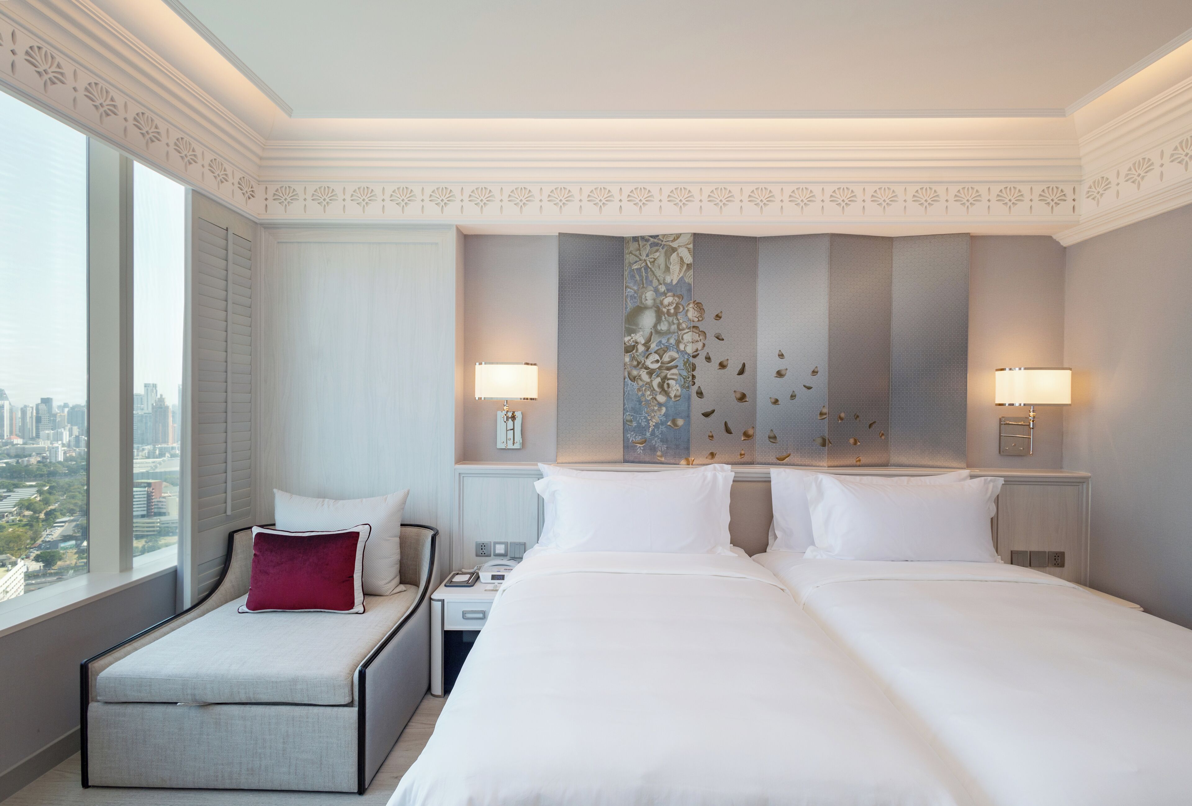 A hotel room with a city view, a bed with white linens, a red accent pillow, two bedside tables with lamps, and a decorative wall panel.